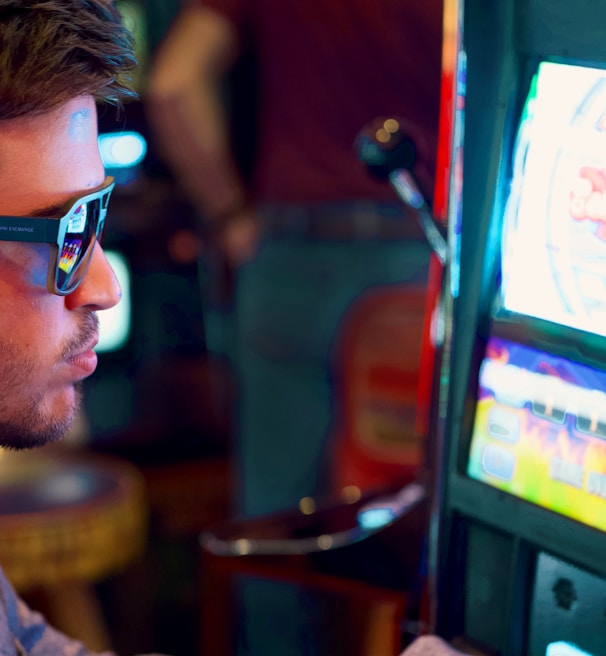 A man wearing sunglasses is intently focused on a slot machine, with colorful lights and graphics illuminating the screen. The scene is set indoors, likely in a casino or gaming area. His attire includes a patterned shirt and a blazer.