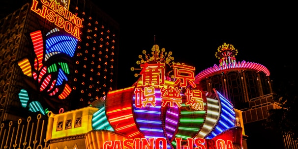 Bright neon lights and colorful signage adorn the exterior of Casino Lisboa, creating a vibrant and lively atmosphere. The façade features bold, multicolored designs with a mix of English and Chinese characters prominently displayed. The lights form intricate patterns and shapes, contributing to the energetic ambiance of the scene.