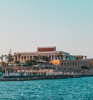 A seaside view of a large building labeled as a casino, situated on a rocky shore. The building is surrounded by palm trees and other smaller structures. The calm sea water reflects the sky, and there are several closed umbrellas lined along the waterfront.